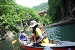 A woman, a girl, and a dog are canoeing on a calm river surrounded by lush green trees and a red bridge in the background.