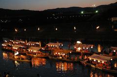 Several boats adorned with lights glide on the water at night, reflecting the illumination against the dark backdrop of hills.