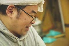 A craftsman in a grey hoodie and a white towel on his head, wearing glasses, smiles while working on tatami mats.