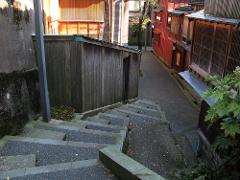 A narrow alleyway descends between traditional wooden buildings in Kanazawa, lined with stone steps and lush greenery.