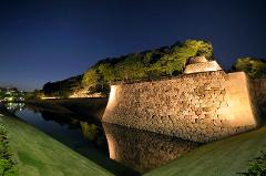 Illuminated stone walls and lush trees surround a tranquil moat reflecting the night sky, hinting at Kanazawa's historical samurai culture.