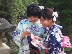 Two women wearing kimonos are looking at a smartphone and a fan with fireworks printed on it, smiling in an outdoor setting.