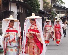 A group of four young women in traditional Japanese attire, including straw hats and white veils, are walking down a street, with a woman holding a baby in the background.