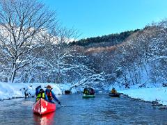 A group of people are canoeing down a river surrounded by snow-covered trees and a clear blue sky.