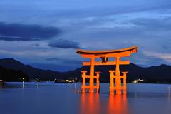The image shows the illuminated orange torii gate of Itsukushima Shrine in Miyajima, Japan at dusk, with mountains and dark clouds in the background.