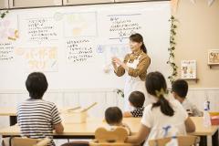 A teacher in a brown shirt and white apron instructs children at a table in a bright classroom with whiteboard instructions and decorations.