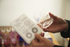 A person's hands hold a piece of paper with instructions for crafting a "Sunflower" pattern and a clear glass with a similar etched design.