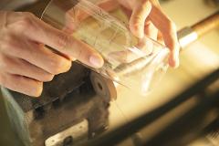 A craftsman's hands hold a clear glass tumbler engraved with delicate wheat stalks while it rests on a grinding wheel.