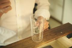 A clear glass with a wheat stalk design sits on a wooden surface, with a person in a white shirt blurred in the background.