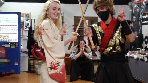 Três pessoas posam com espadas de madeira, uma mulher de quimono, um homem com traje ninja e outro homem sentado ao fundo.