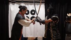 Duas pessoas vestidas com roupas tradicionais japonesas estão envolvidas em uma luta de espadas, uma de frente para a outra com suas espadas cruzadas.