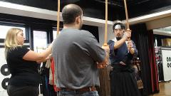 Um homem de camiseta cinza e jeans, uma mulher loira e um homem em trajes tradicionais japoneses empunhando espadas de madeira estão em uma sala.