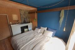 A cozy bedroom features a bed with light-colored linens and pillows, set against a backdrop of blue and textured walls, with decorative elements like plants and a stone fireplace adding warmth.