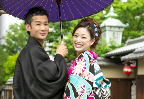 A happy Japanese couple in traditional clothing shares an umbrella and smiles at the camera.